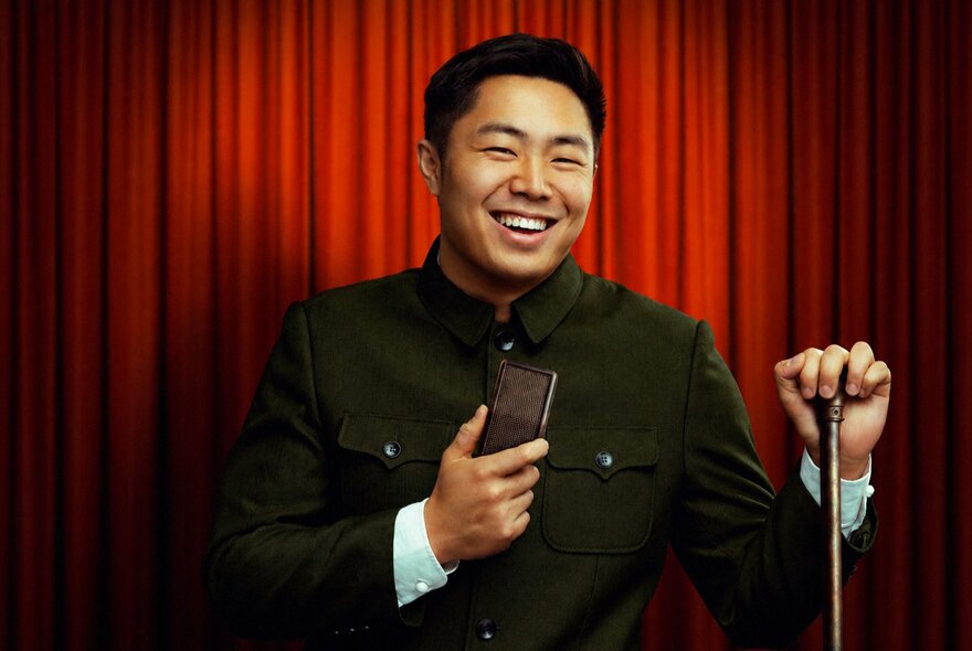 Comedian Nathan Chin on stage with an old-fashioned microphone, in front of a red stage curtain.