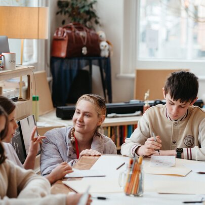 A teacher bobs down next to a seated students writing at a table in a studio workshop and listens intently.