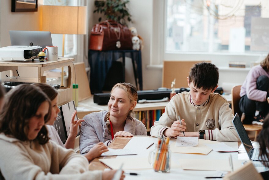 A teacher bobs down next to a seated students writing at a table in a studio workshop and listens intently.