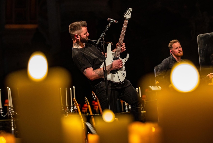 A man playing electric guitar on a candlelit stage, seen through blurred candles in the foreground.