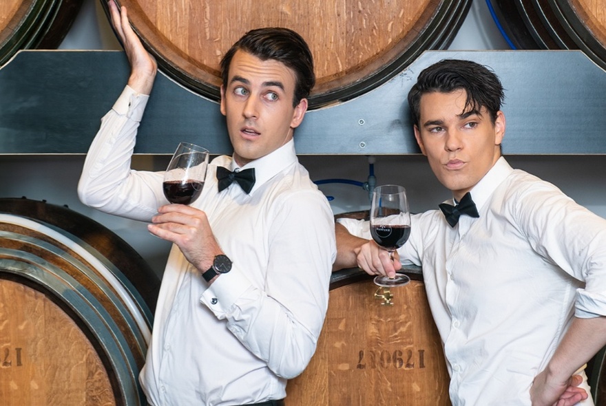 Two men dressed formally with white shirts and black bowties holding glasses of red wine and leaning on wooden barrels.