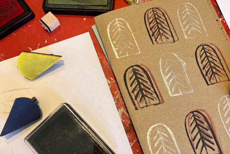 Stamped designs of a Christmas tree outline in an arch shape in white and purple ink, on a piece of brown card, the carved stamps and ink pad resting on the table alongside. 