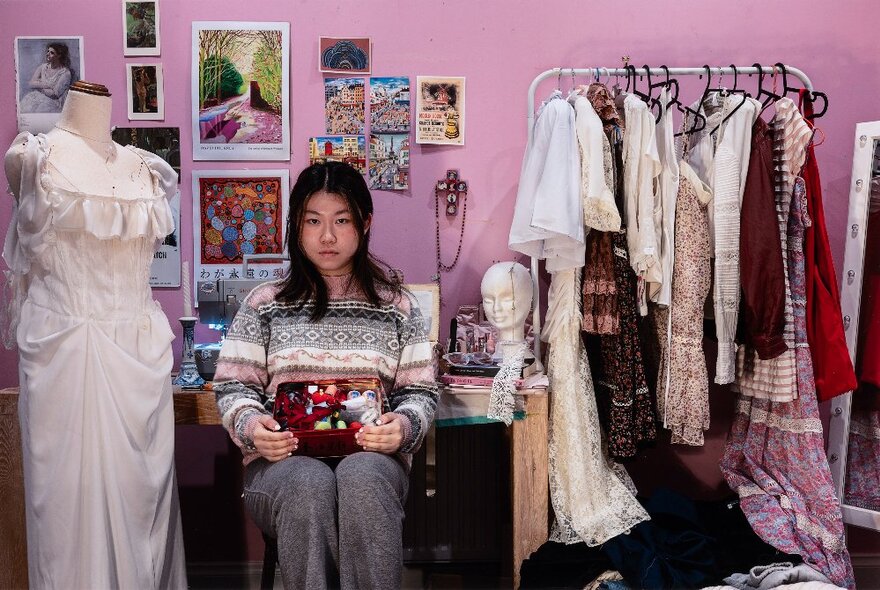 Young woman sitting facing the viewer with a box of sewing paraphernalia on her knee, behind her is a sewing machine, and there are drawings and photos pinned to the wall, and a rack of garments to her left.