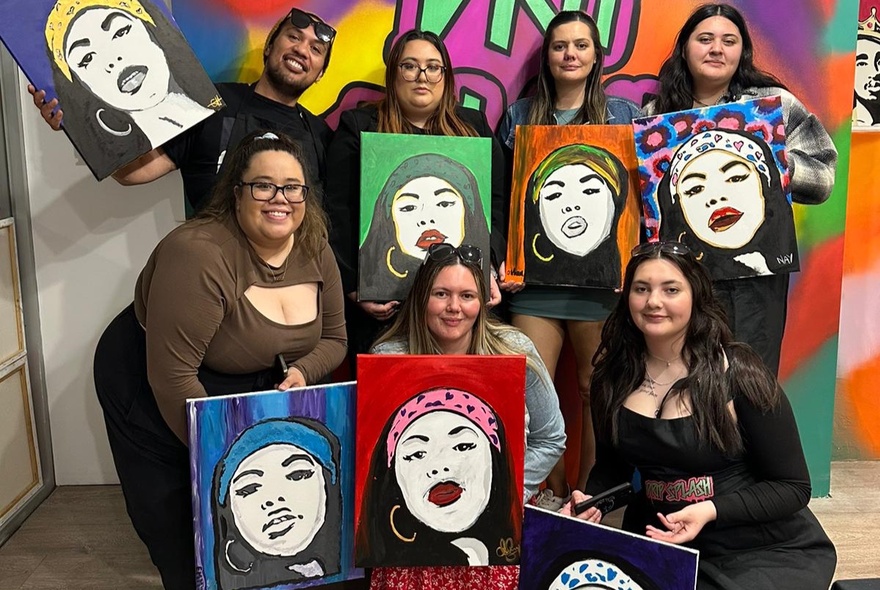 Two rows of people smiling and holding up canvases they have painted in a room with large, wall murals.