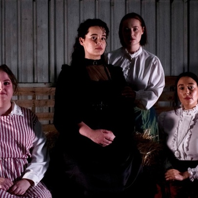 Actors dressed in 19th-century dresses, posing with hands clenched.