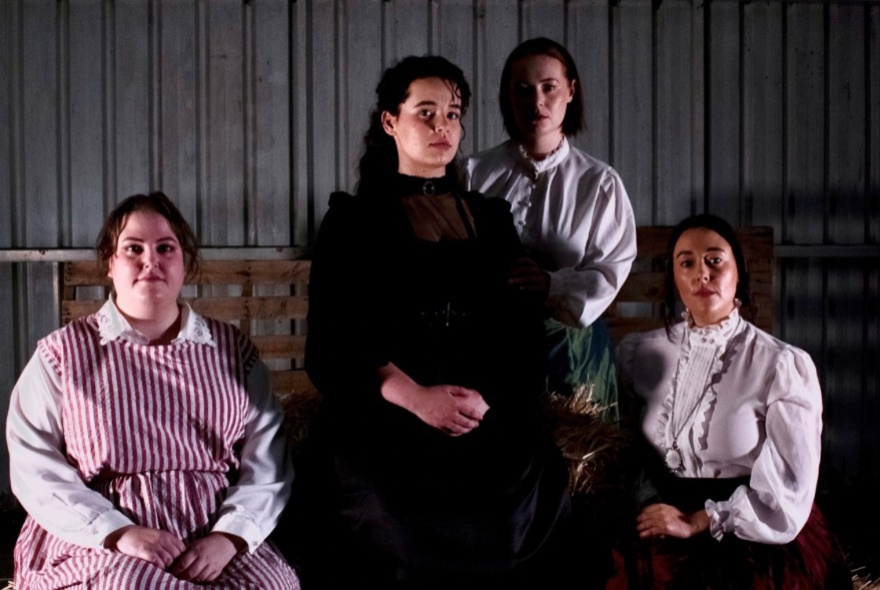 Actors dressed in 19th-century dresses, posing with hands clenched.