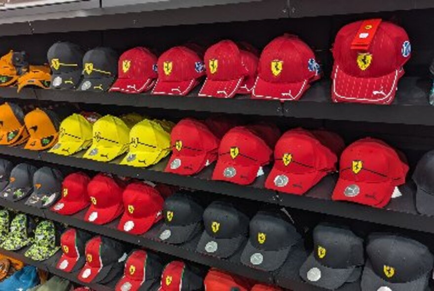 Many F1 branded racing caps in different team colours, on display on shelves in a shop.