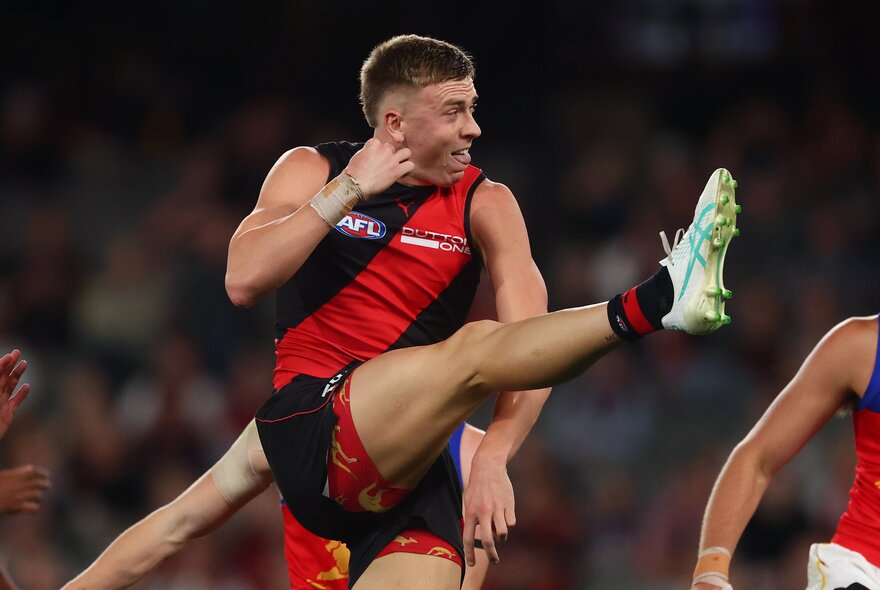 An Essendon AFL football player with his leg raised high in the air, having just kicked the football