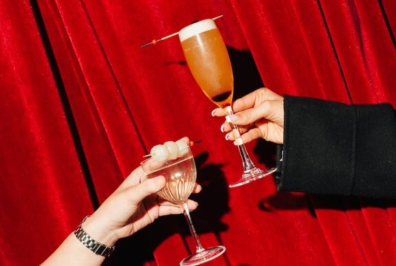 Two hands raising glasses in a toast in front of a red velvet curtain. 