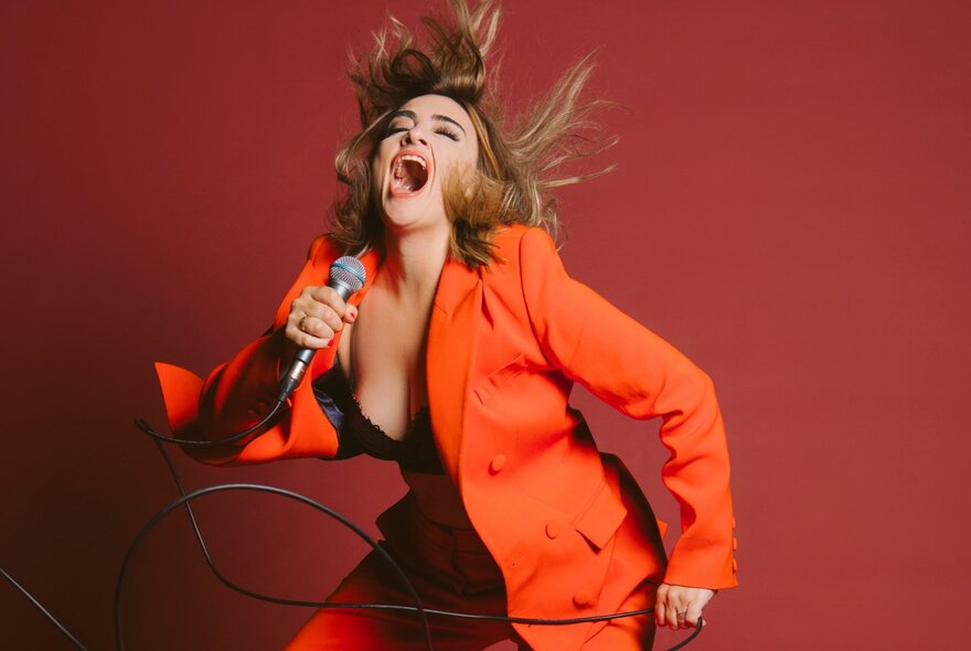 Performer in an orange blazer and trousers, singing into a microphone she holds, throwing her head and hair back, set against a red background.