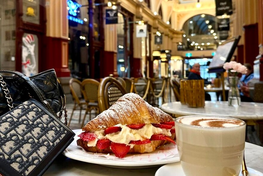 A strawberry and cream croissant on a plate next to a latte and a patterned handbag on a cafe table, with an ornate shopping arcade walkway softly blurred in the background.