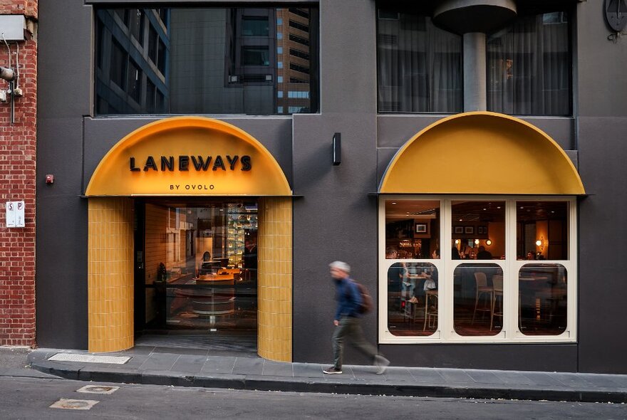 Exterior of Laneways by Ovolo, a dark grey buildign with banana yellow accents, a man walking past slightly uphill. 