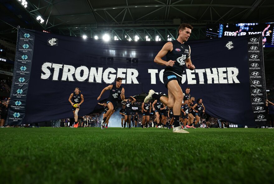Carlton AFL football players running through their team's banner which reads 'STRONGER TOGETHER' and onto the field at the start of the match. 