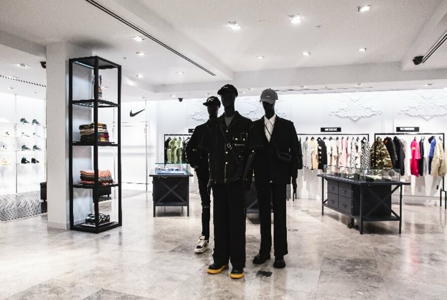 Interior of Harrolds designer fashion store, large open space, white walls, ceiling and tiled floor, with three mannequins in black clothing and a rack of clothes against the rear wall.