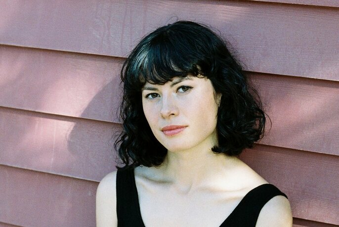 Head and shoulders of a pale young woman with shoulder length dark hair, looking directly at the viewer while leaning against dusty pink coloured weatherboards.