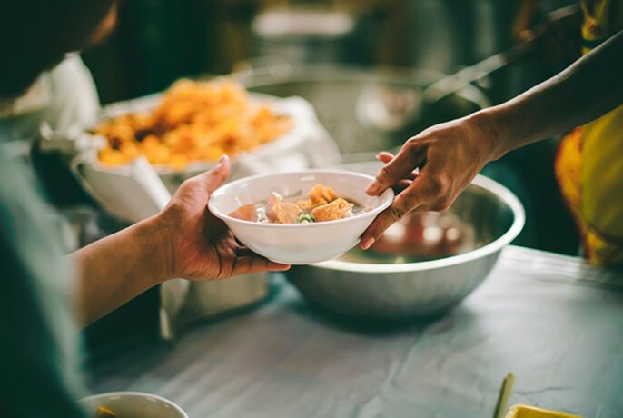 A person handing a bowl of food to another person with large bowls blurred in the background. 