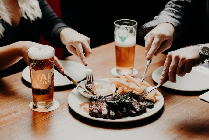 Two people cutting into a plate of meat and chips with beer.
