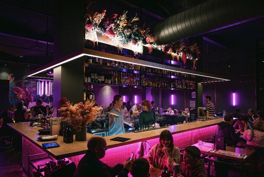 Bar service area in centre of a restaurant, illuminated with pink light, dining tables with patrons eating and drinking around it.