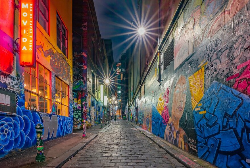 A cobblestone laneway at night with bright street art murals. 