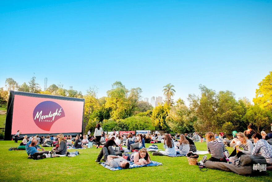 Late afternoon in the Royal Botanic Gardens Moonlight Cinema area with a large movie screen in front of sunlit trees with a crowd of people on picnic blankets on the grass.