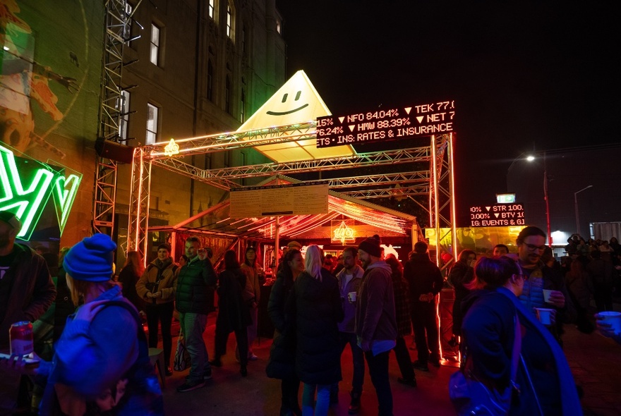 Night scene of an outdoor space filled with people walking around exploring the installations and food and drink stalls around them.