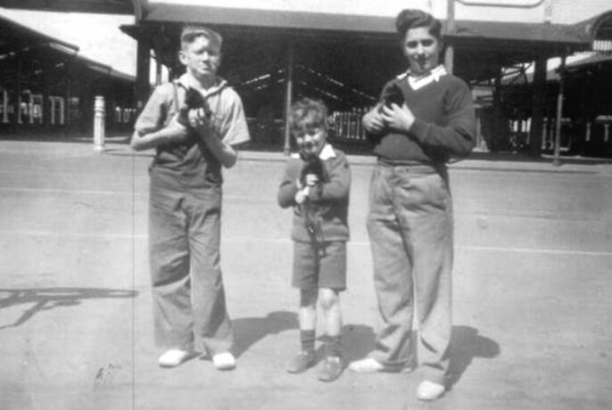 Vintage black and white image of three young boys dressed in 1950s style clothing, all holding either small kittens or rabbits in their arms, close to their chest, in front of a large empty shed.