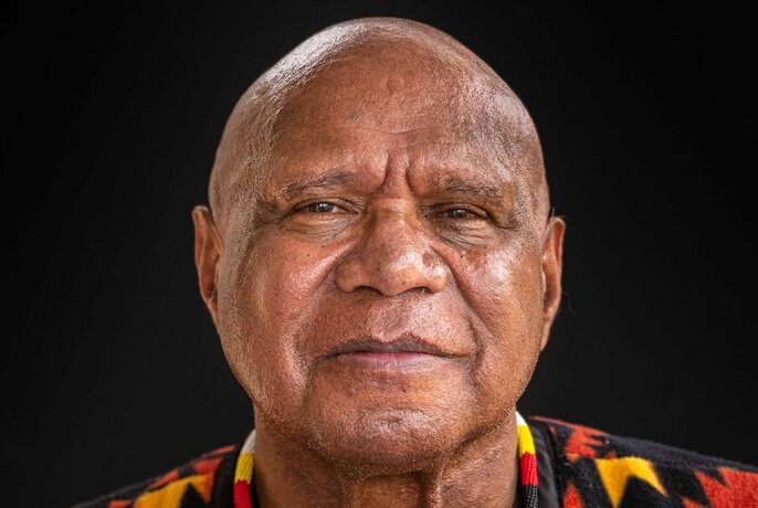 Close up headshot portrait of the singer and musician Archie Roach, a bald man in his 50s, smiling very slightly, a black background behind him. 