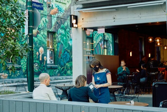 Patrons sitting at footpath tables and chairs, outside the front of Lane's Edge Wine Bar, a painted mural on the laneway wall visible in the background.