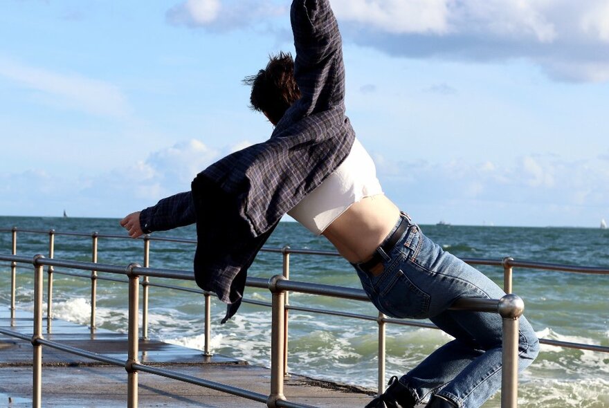A person seated on a railing overlooking the sea, with arms wildly waving in the air and torso bared.