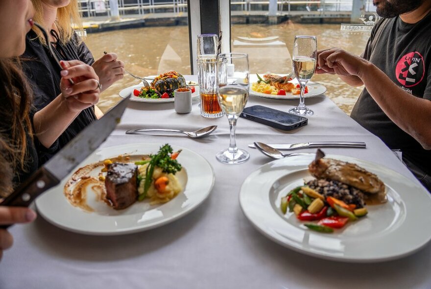 Diners seated at a restaurant table on a floating river cruise restaurant, with plates of delicious food in front of them and the water of the Yarra River in the background.