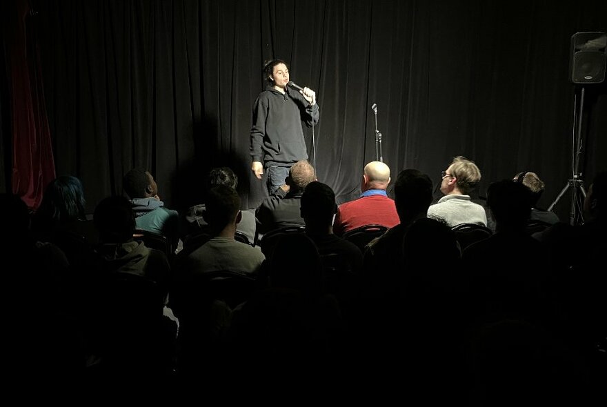 Person in dark clothing holding mic, in front of audience in darkened room.