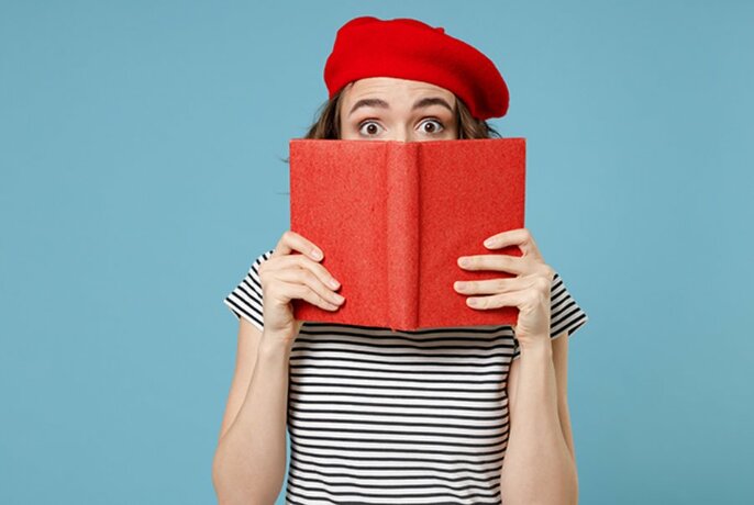 Person wearing a black and white striped t-shirt and red beret, holding up a red book in front of their face, with just their eyes and eyebrows visible above the book.