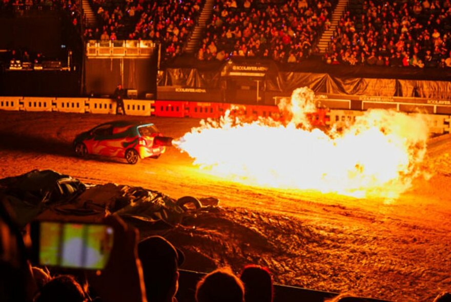 A rally car with a jet of fire coming out of its rear exhaust, driving on a dirt track inside a stadium, with spectators watching.