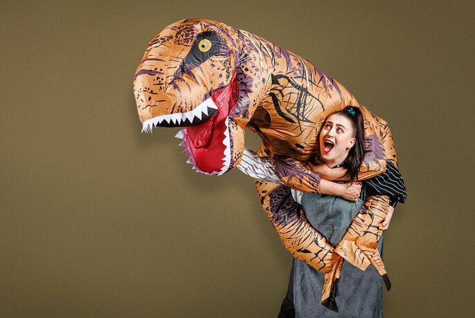 Person in grey shift and dark hair, with mouth open in delight and surprise, with inflatable dinosaur on shoulders.