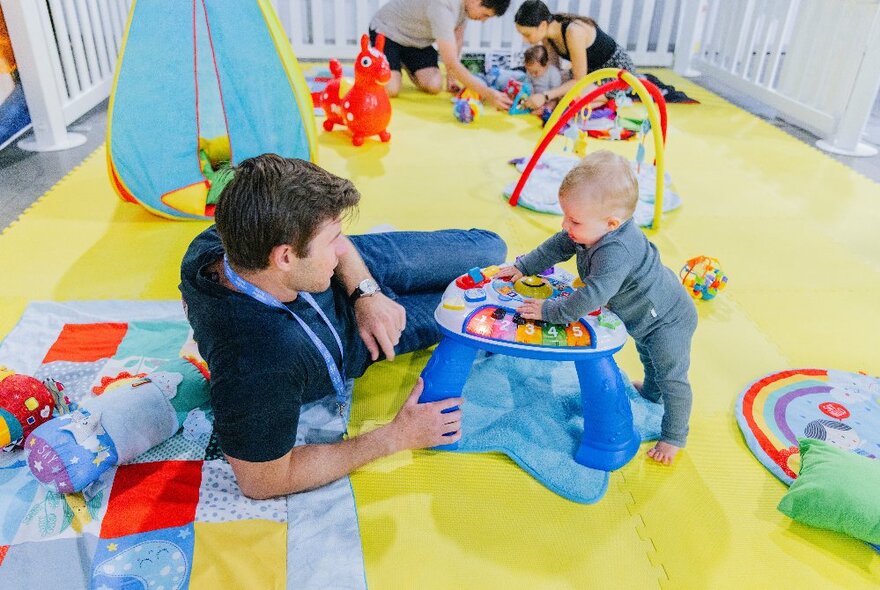 Adult and a baby interacting in a soft play area, with soft rugs and toys strewn around. 