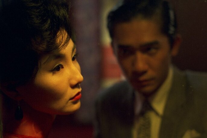 Movie still showing a woman looking wistfully to the side of the frame while a man in the background looks at her.