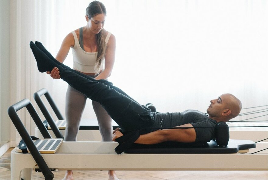A woman helping a man get into position on a pilates reformer.