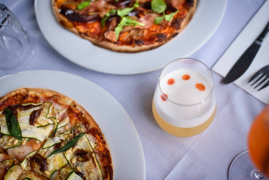 Bird's eye view of two pizzas and a cocktail on a table with white tablecloth.