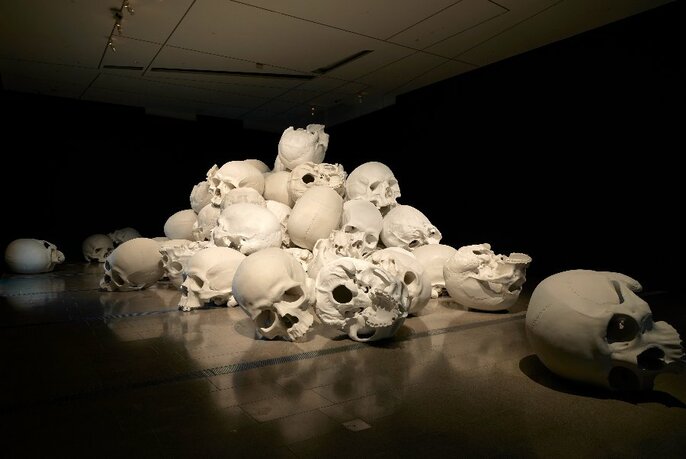 Installation view of white resin casts of a group of oversized human skulls, arranged in close proximity in a pile.