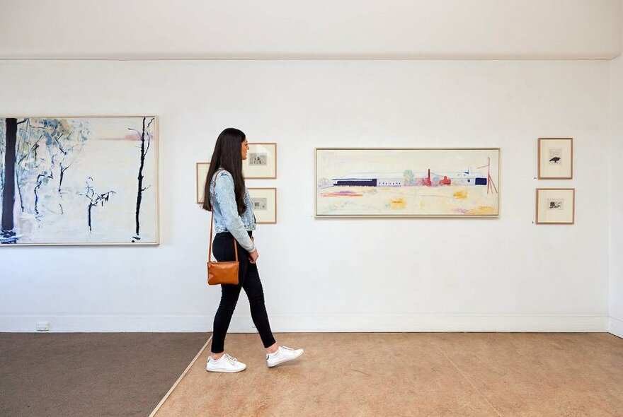 A woman walking through a gallery looking at paintings on the wall.