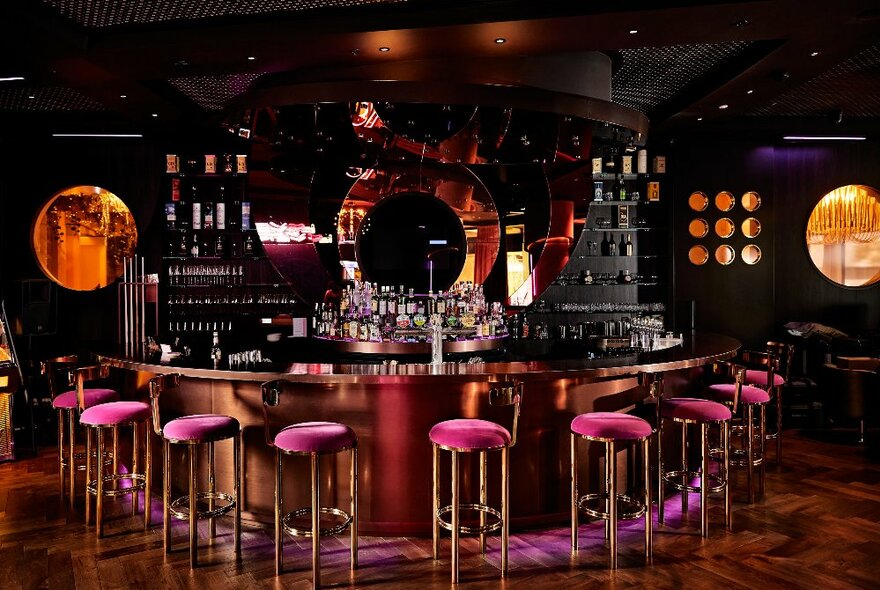 A glamorous cocktail bar with plush pink stools around a semi-circular bar and circular windows. 