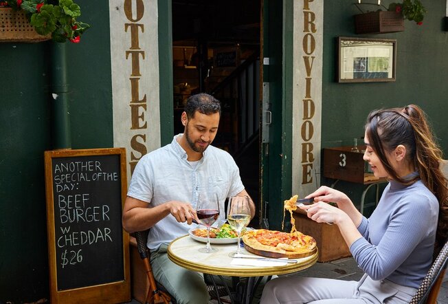 A couple dining on pizza and wine outside a restaurant.
