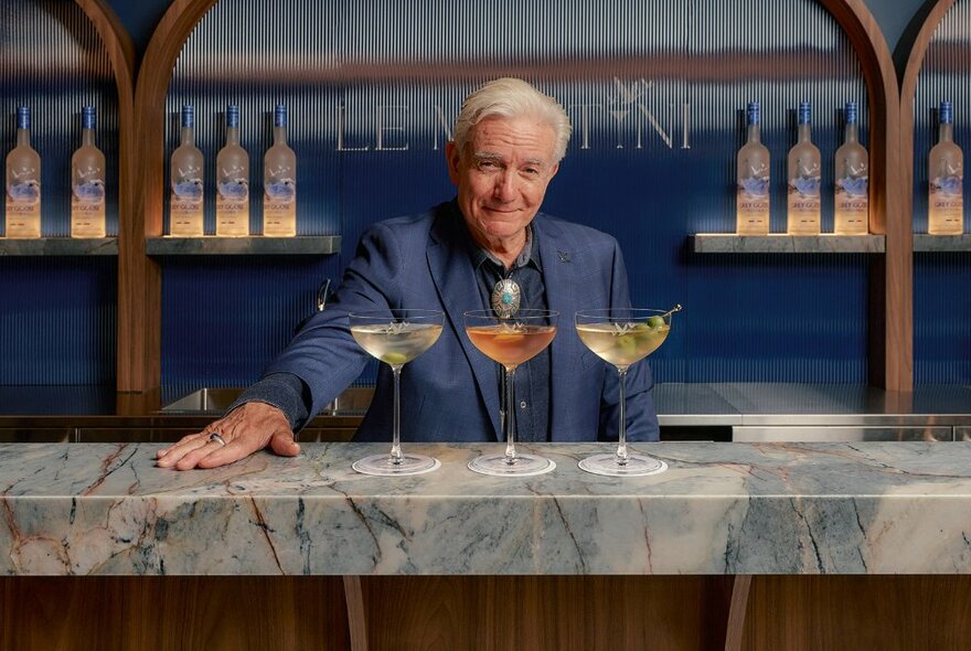 Experienced bartender Dale De Groff standing  behind a bar with three martinis on the marble bar counter in front of him.