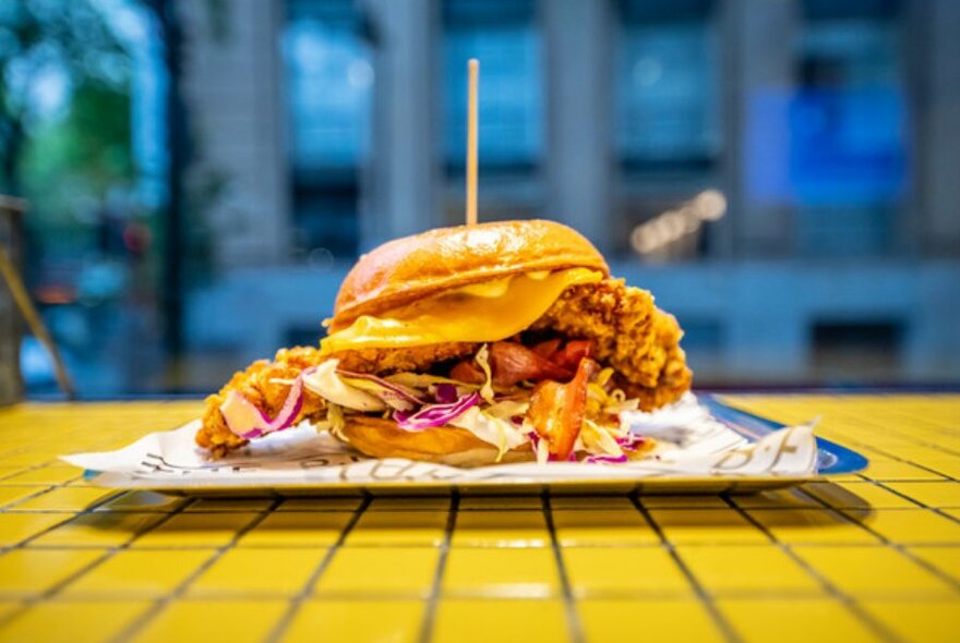 Close up shot of a fried chicken burger with cheese and coleslaw. Burger is on a metal tray placed on a yellow tiled bench top. 