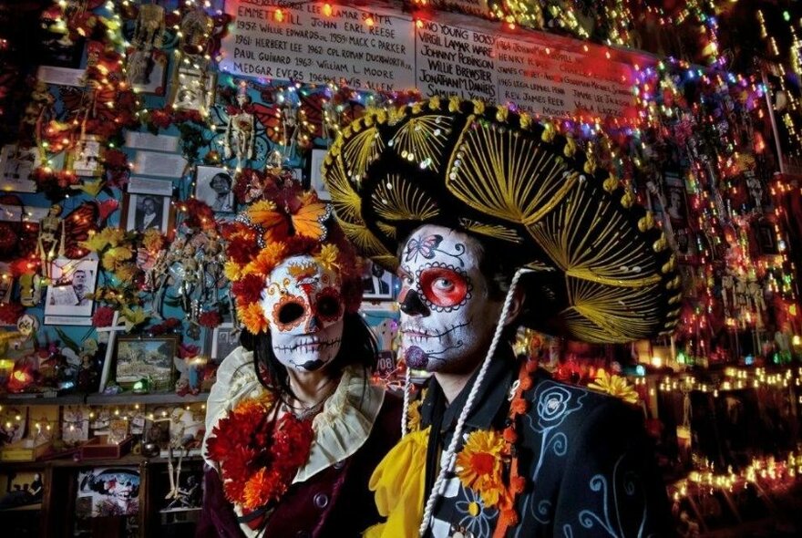 Festival-goers dressed in Mexican Day of the Dead outfits and makeup, walking past candle-lit stalls.