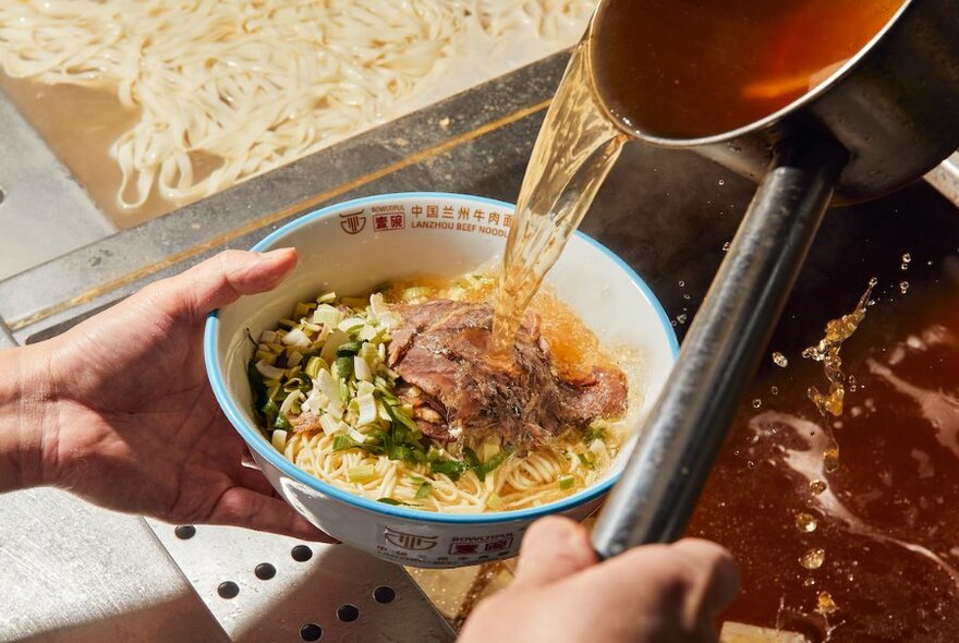 Pouring stock over a large bowl of noodles and and beef. 