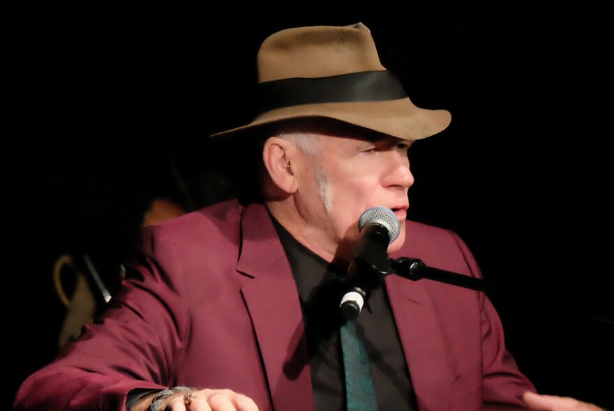 Stewart D'Arrietta sitting and performing in a burgundy jacket and brown hat.