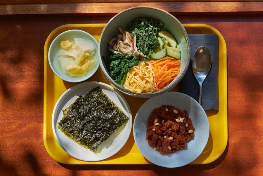 Looking down on a bowl of bibimbap with condiments on a yellow serving tray.