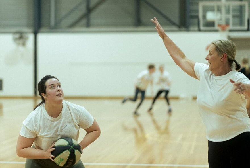 Two women playing basketball inside a basketball stadium, with other women in the background.