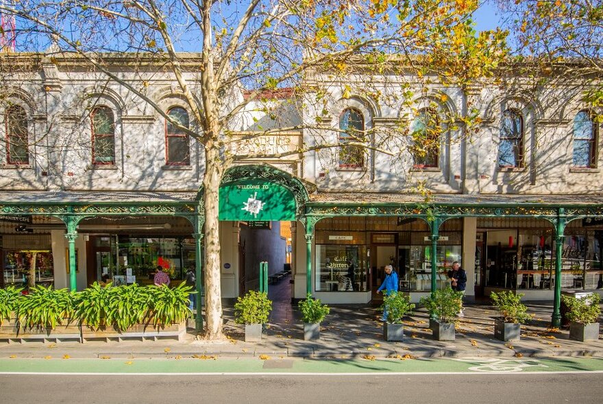 a man walking in front of a strip of cafes along a leafy street